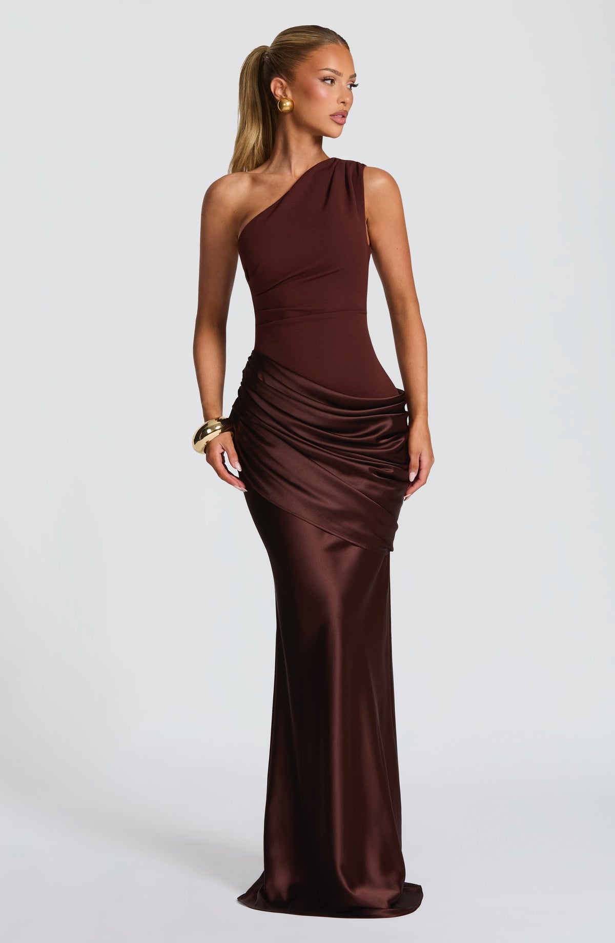 Brown one-shoulder satin gown with ruched hip drape, worn with a gold cuff and earrings against a plain grey backdrop. 14
