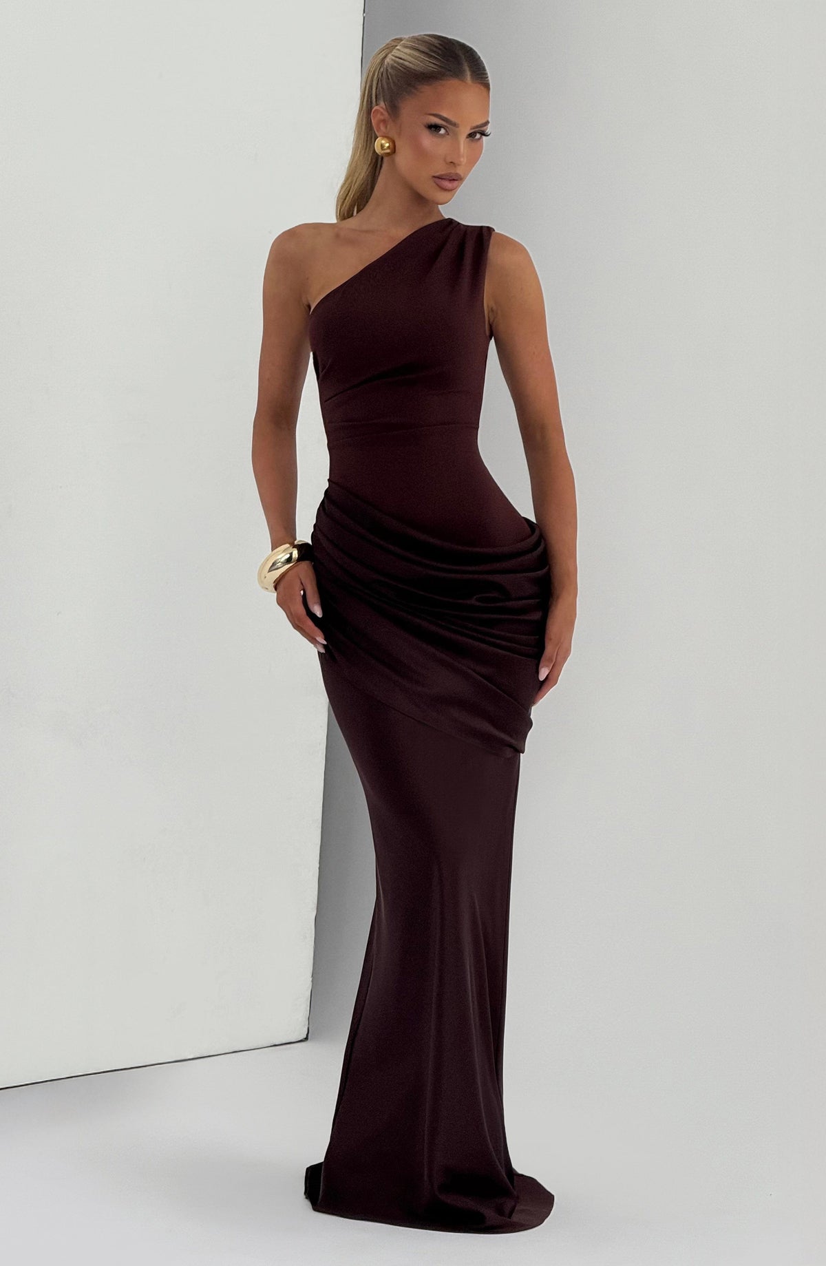Brown one-shoulder maxi dress with draped hip panel, styled with gold earrings and a chunky gold cuff. 9