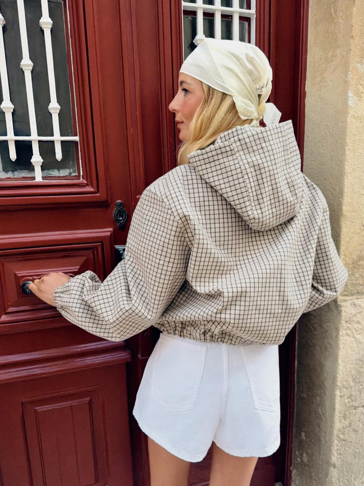 Back view of the model reaching for a door handle, showing the checked hood, elasticated cuffs and gathered hem. 4