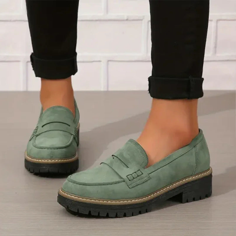 Green suede-look penny loafers with chunky black treaded soles, worn with rolled black trousers against a white brick wall. 1