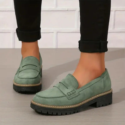 Green suede-look penny loafers with chunky black treaded soles, worn with rolled black trousers against a white brick wall. 1