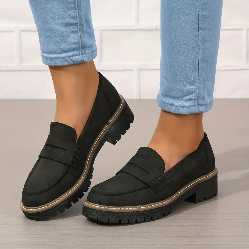 Black suede-look penny loafers with chunky treaded soles, worn with cuffed blue jeans against a white brick wall. 6