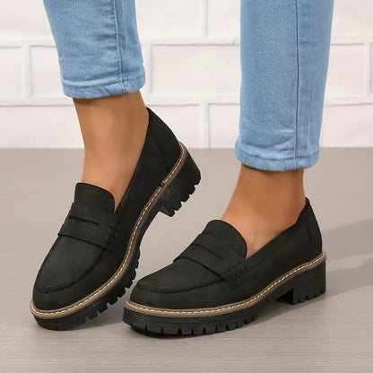 Black suede-look penny loafers with chunky treaded soles, worn with cuffed blue jeans against a white brick wall. 6