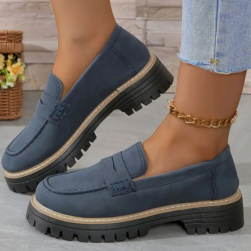 Navy blue suede-look loafers with a tan welt and chunky black platform sole, worn with a gold chain anklet. 16