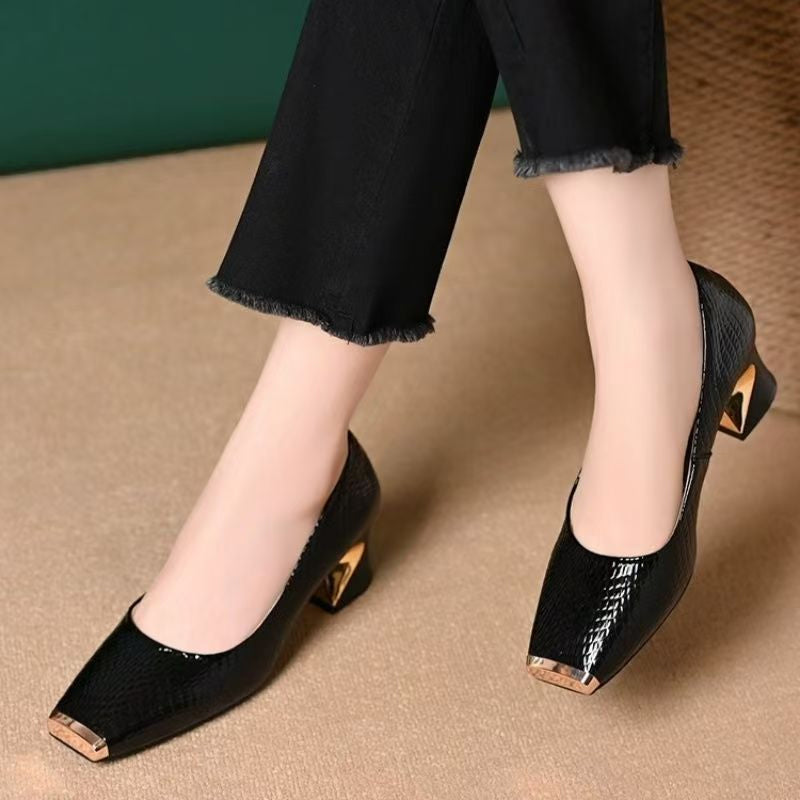Black glossy snake-effect pumps with gold metal toe caps and sculpted gold-accent heels, worn with frayed black cropped trousers on a beige floor. 1