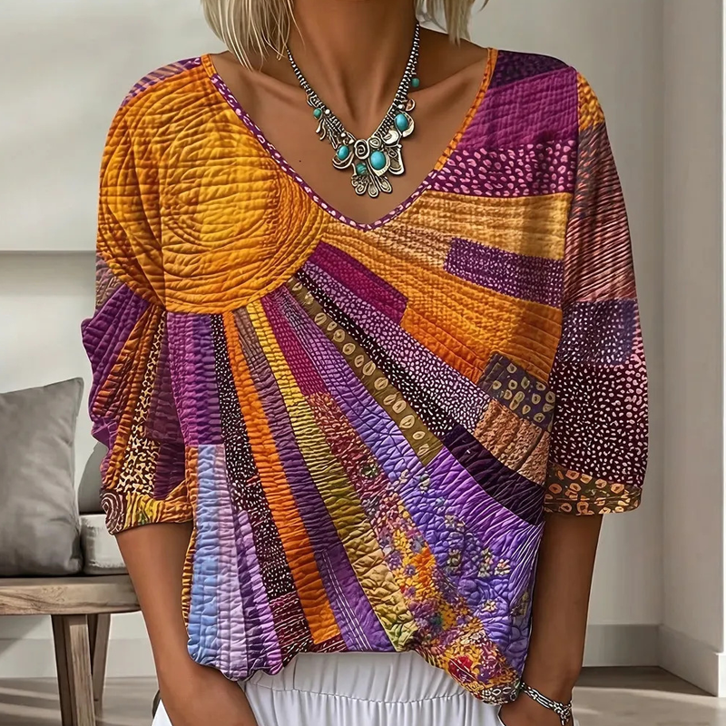 Close-up of the sunburst patchwork blouse in orange, purple and magenta tones, worn with white trousers and a turquoise statement necklace. 2