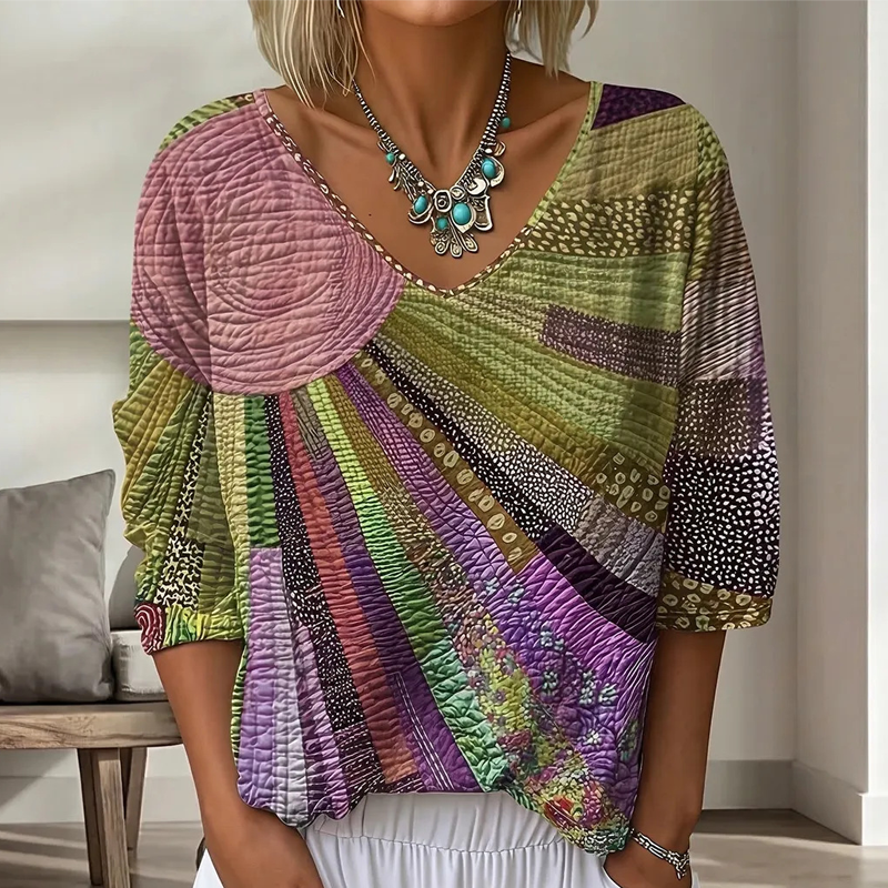 Close-up of the sunburst patchwork blouse in olive green, pink and purple tones, with a quilted texture and contrast trim along the V-neckline. 4