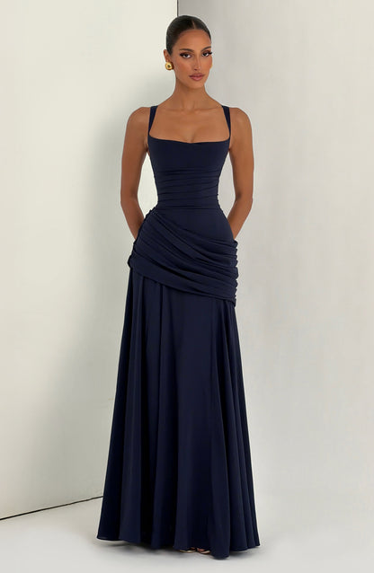 Navy maxi dress worn by a model against a light studio corner, front view with hands behind her back and the skirt flowing. 13
