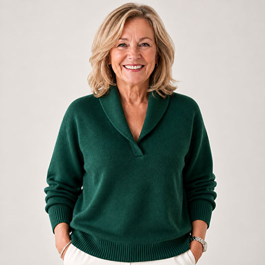 Smiling woman wearing the knitted jumper in dark green, hands in the pockets of white trousers, shown from the front. 1
