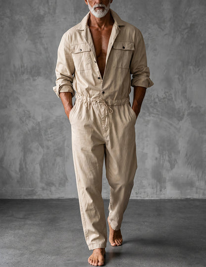 Barefoot model in a beige long-sleeved jumpsuit with rolled cuffs, walking against a grey textured wall 1
