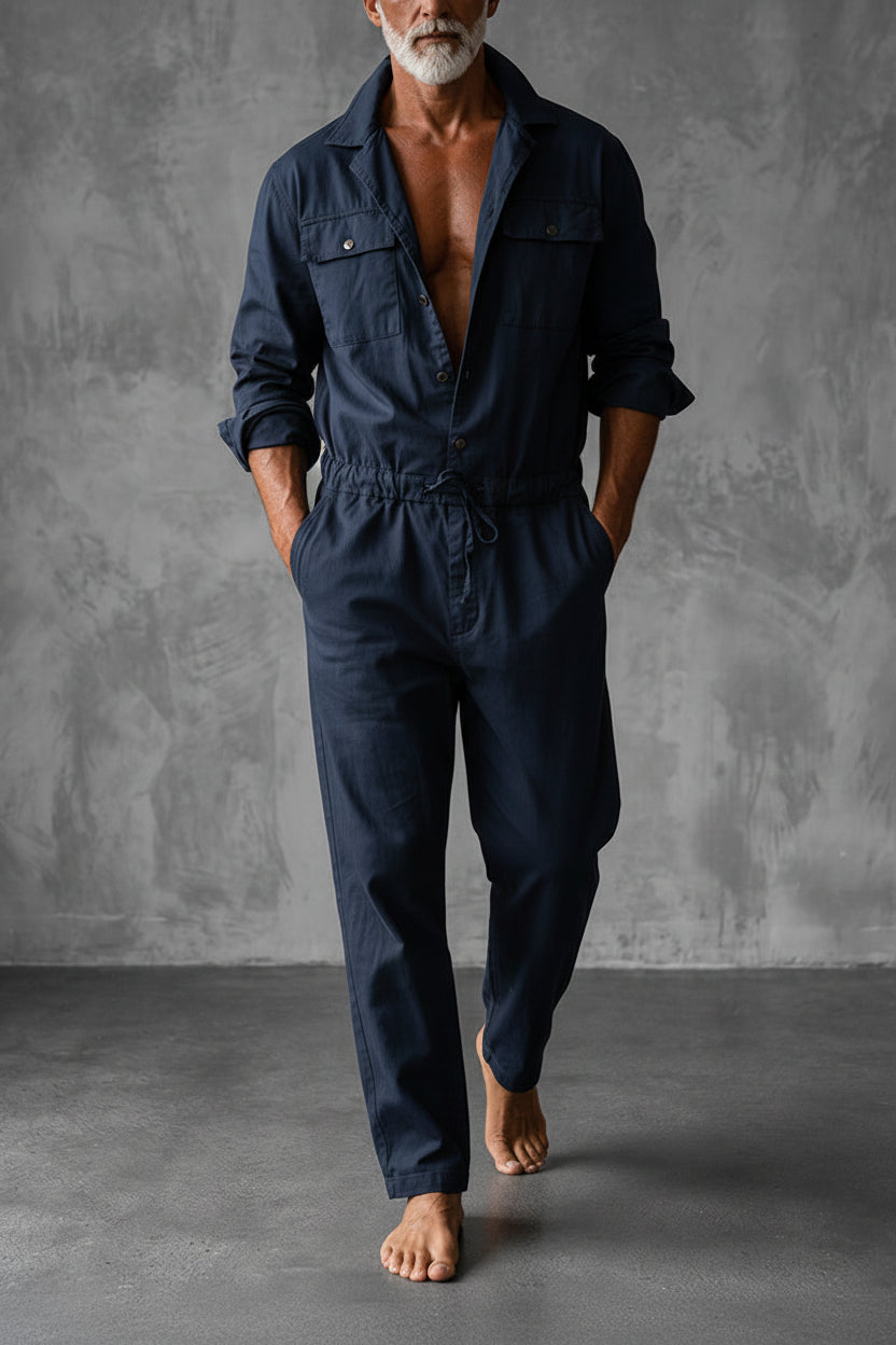 Same jumpsuit in navy, worn open at the chest by a barefoot model against a grey wall 2