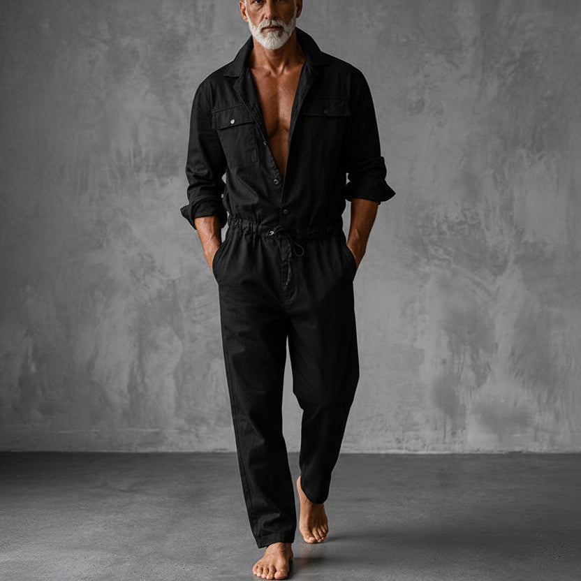 Black version of the jumpsuit, worn by a barefoot model with hands in pockets, grey wall behind 3