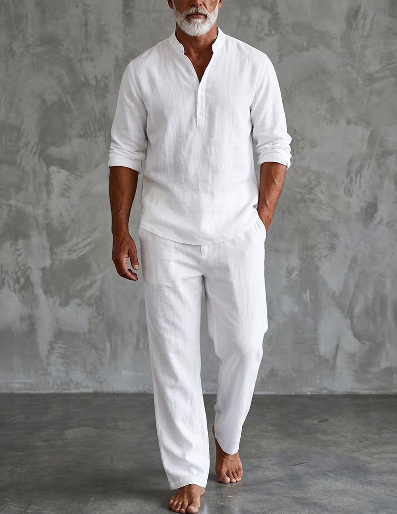 Barefoot bearded man in the white set, sleeves rolled, one hand in pocket, standing against a grey textured studio wall. 1