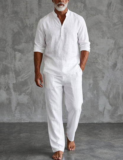 Barefoot bearded man in the white set, sleeves rolled, one hand in pocket, standing against a grey textured studio wall. 1