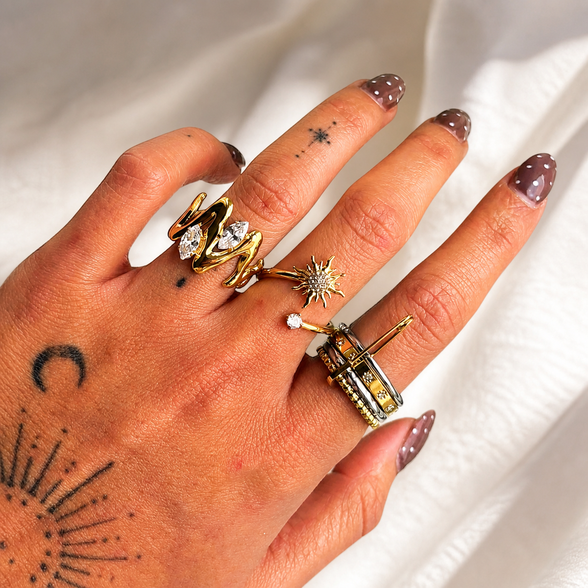 Hand with tattoos and brown polished nails wearing the wavy gold-toned ring on one finger, stacked with other rings. 3