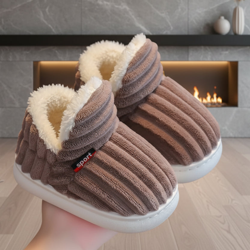 Pair of brown ribbed plush slippers with white fleece lining, held in a hand in front of a fireplace. 1