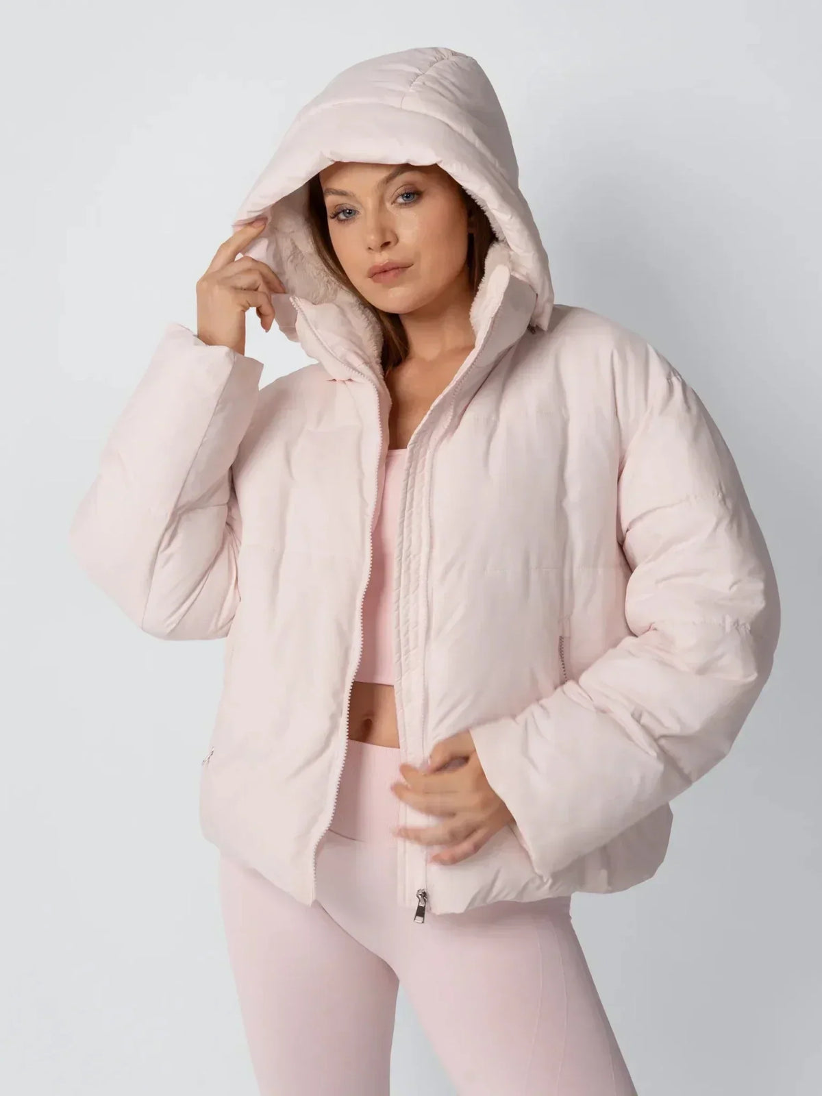 Model in a pale pink puffer jacket with the hood up, touching the hood edge and showing the fleece lining 6
