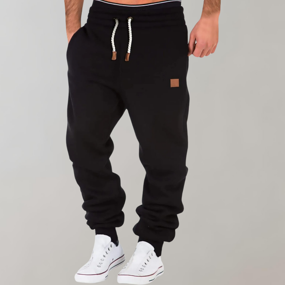 Model wearing black joggers with white drawstring, ribbed ankle cuffs and white trainers, one hand in pocket 1