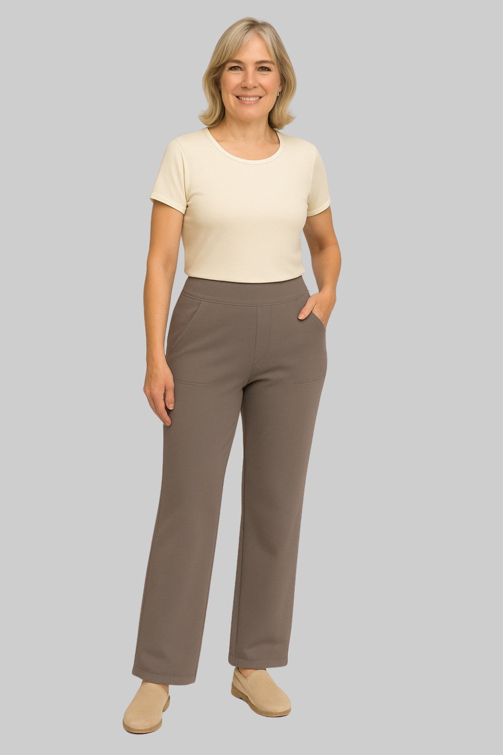 Smiling woman wears the taupe straight-leg trousers with a cream short-sleeved top and beige slip-on shoes, one hand in a pocket. 2