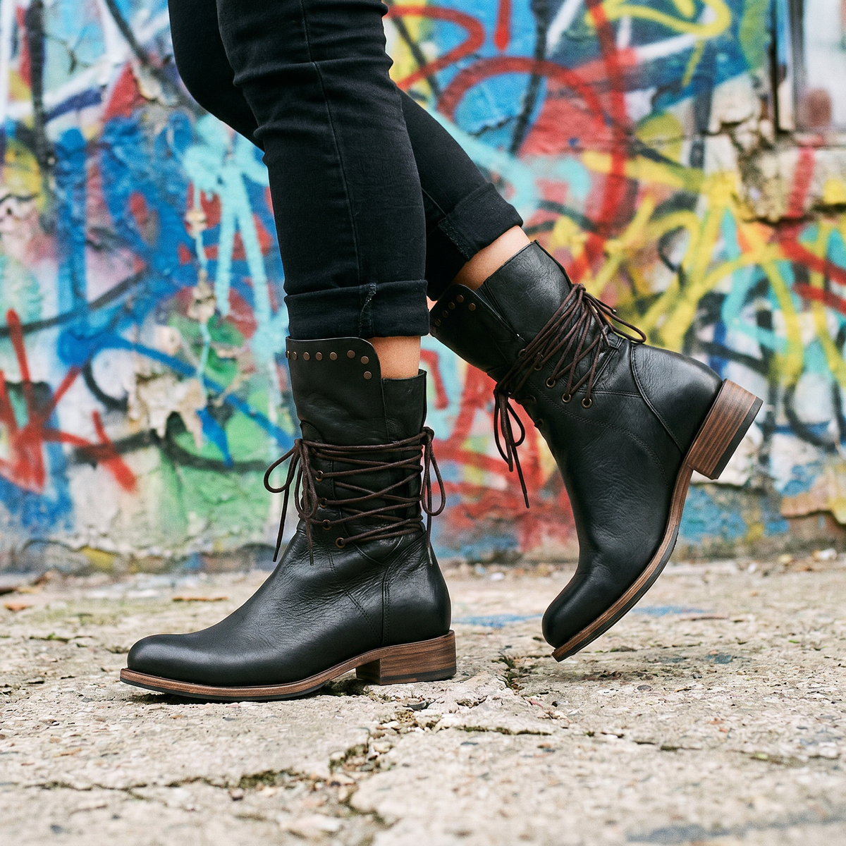Side view of black lace-up boots with black jeans, mid-step in front of a colourful graffiti wall. 19