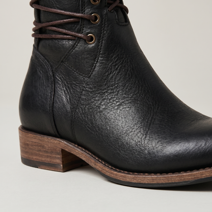 Close-up of a black leather boot's grained leather, brass eyelets, stitched welt and low stacked wooden heel. 6