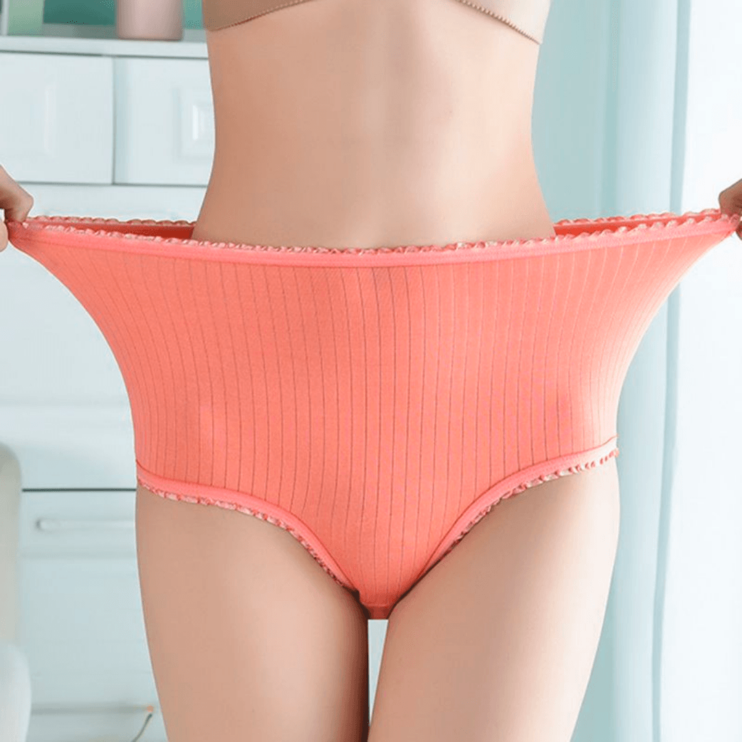 Model stretching the waistband of coral ribbed high-waisted briefs outwards to show how elastic the fabric is, with frilly trim. 1