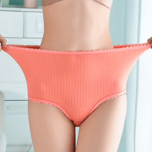 Model stretching the waistband of coral ribbed high-waisted briefs outwards to show how elastic the fabric is, with frilly trim. 1