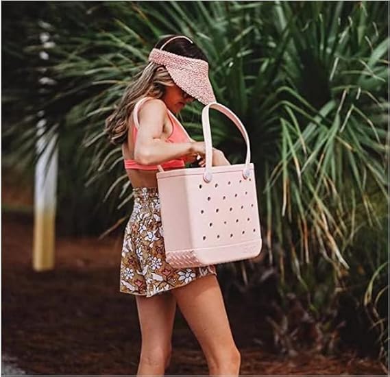 Woman in a straw visor and floral shorts reaching into a pale pink perforated tote carried by its handles outdoors. 1