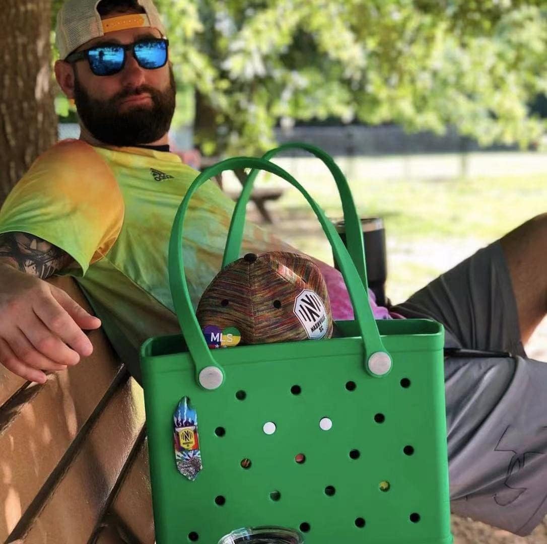 Bearded man in sunglasses sitting on a bench beside a green tote holding a cap, with small charms attached to the holes. 13