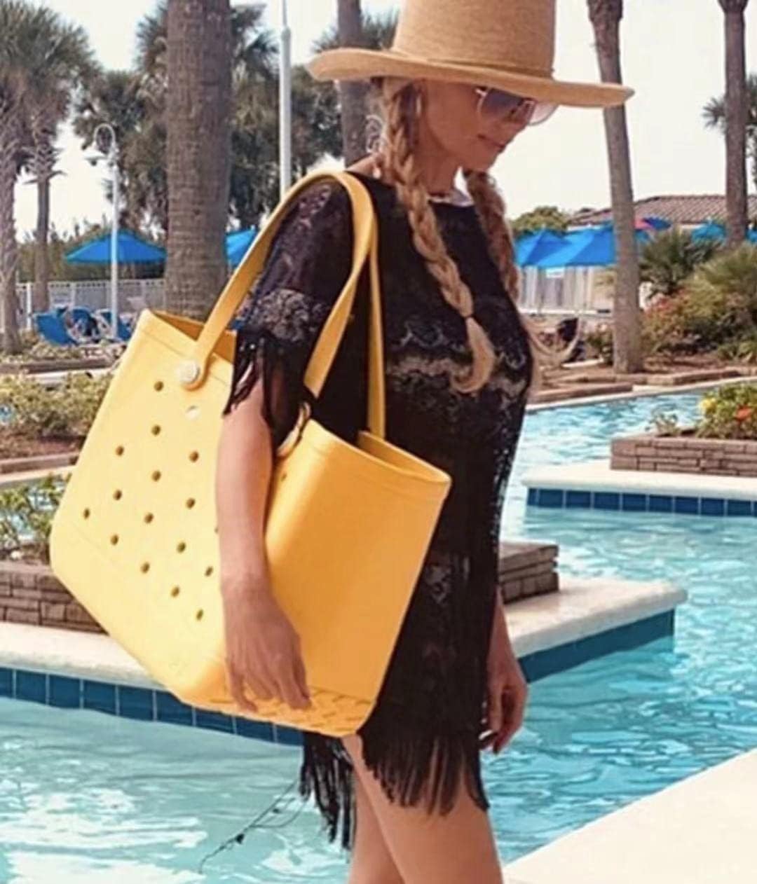 Woman in a black lace cover-up and straw hat carrying a yellow perforated tote over her shoulder beside a pool. 2