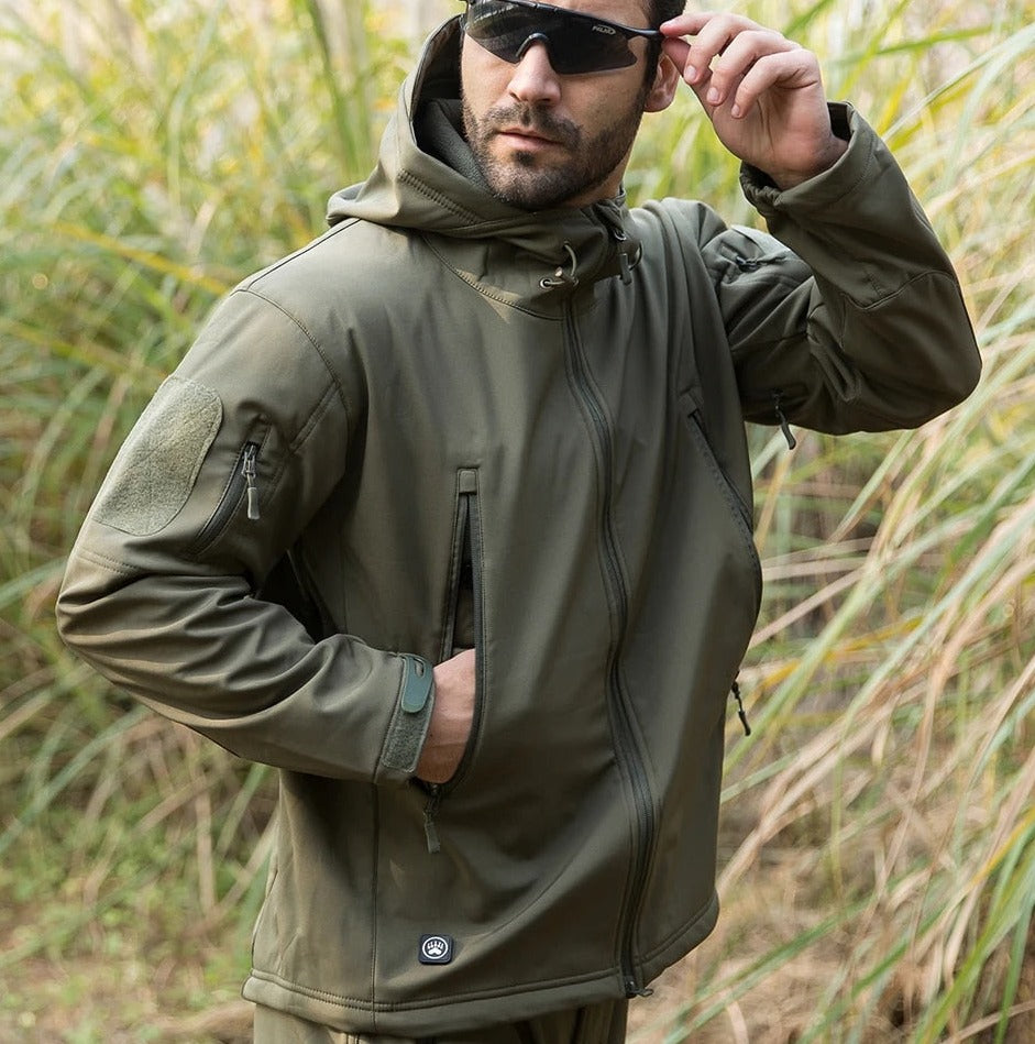 Man in sunglasses wearing the olive green jacket outdoors among tall grass, one hand in a pocket and the other touching his glasses. 11