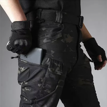 Close-up of dark camouflage trousers worn with a belt and gloves, with a phone being slipped into the zipped thigh pocket. 19
