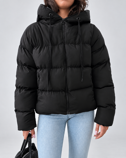 Front view of the black puffer jacket zipped up, showing hood drawstrings, horizontal quilting and zipped side pockets, worn with light jeans. 2